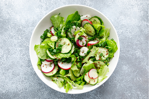 Top-down view of a fresh green salad with sliced cucumbers, radishes, green onions, dill, and flax seeds in a white bowl on a grey stone surface
